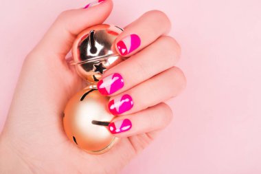 Parlak manikürlü iki kadın eli pembe arka planda izole edilmiş altın şıngırtı çanlarını tutuyor..