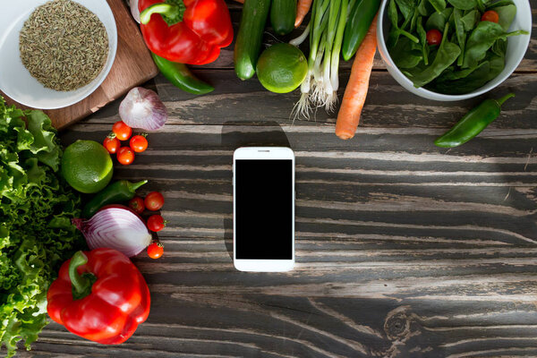 Fresh organic vegetables and smartphone in the center on wooden background, concept of digital recipe book and shopping list.