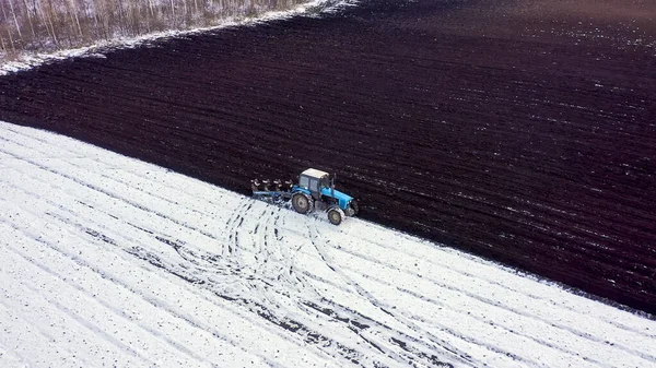 青いトラクターは雪で覆われたフィールドを耕す トラクターの後ろに黒い大地です ロシア ウラル 空中ビュー — ストック写真