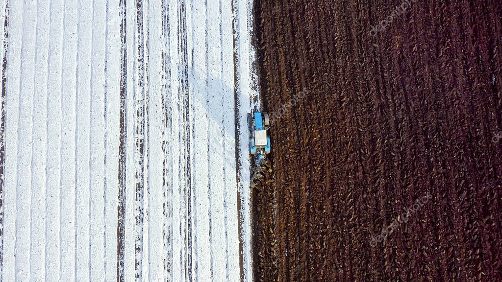 A blue tractor plows a field covered with snow. Behind the tractor is black earth. Russia, Ural, Aerial View, HEAD OVER SHOT  