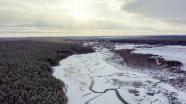 Karla kaplı küçük bir nehri olan bataklıklar. Arka plandaki köy. Bulutlar ve güneş. Ağaçlarda kar, hava manzarası  