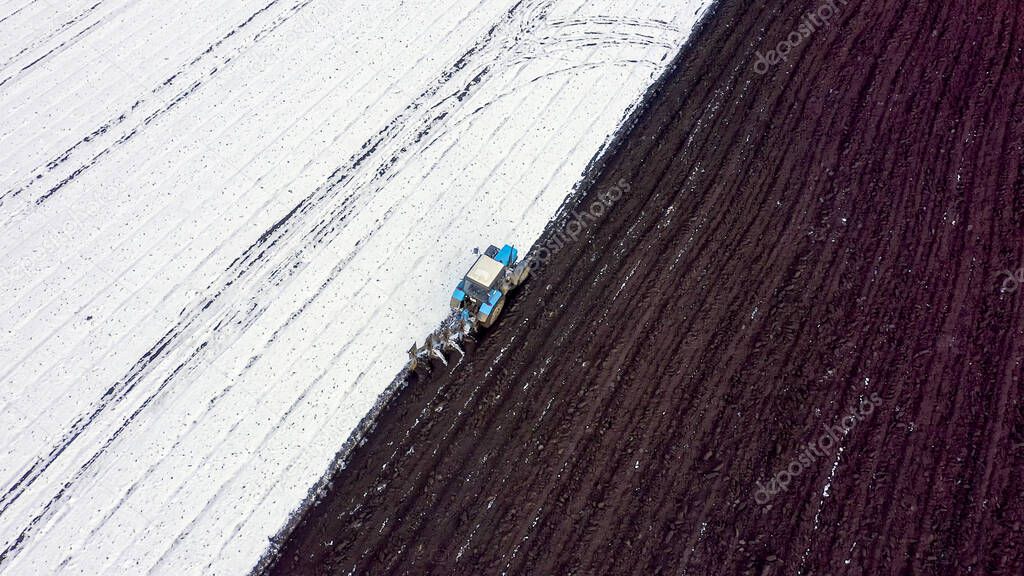 A blue tractor plows a field covered with snow. Behind the tractor is black earth. The edge of the field. Russia, Ural, Aerial View  