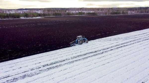 青いトラクターは雪で覆われたフィールドを耕す トラクターの後ろに黒い大地です ロシア ウラル 空中ビュー — ストック写真