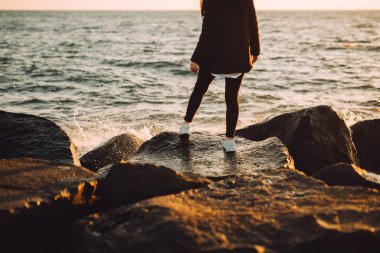 Genç kız deniz kenarında taşlar üzerinde duruyor. Güzel gün batımı arkadan sanki