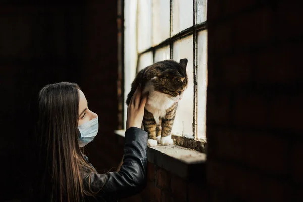 Young woman with her sad cat, sitting at home during coronavirus quarantine. They are home near vintage window. Sad cat looking out the window. Isolation at home to prevent virus pandemic.