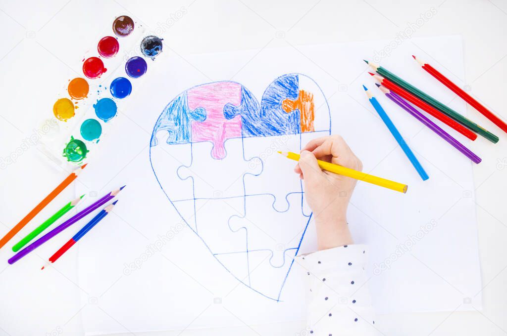 A child is drawing colorful heart with jigsaw puzzle on white background as a symbol of autism, with colorful pencils and watercolors.