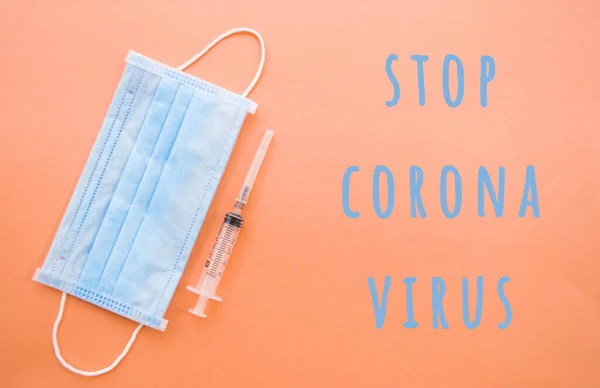 A syringe with a needle and a blue face mask on orange color background. Stop coronavirus concept. Protect yourself.