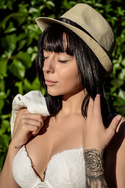 Retrato de menina em roupa interior leve e chapéu e camisa branca em sua mão sobre o fundo de plantas verdes — Fotografia de Stock