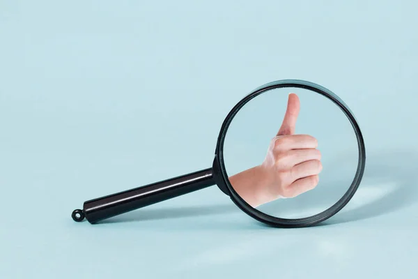 Closeup of female hand showing thumbs up throw Magnifying glass, on a pastel blue background — Stock Photo, Image
