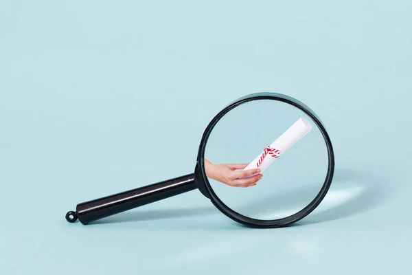 A magnifying glass with a hand handing out a degree or a certficate under it. — Stock Photo, Image