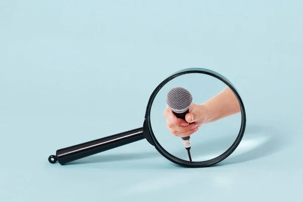 A magnifying glass over a blue background focusing on a hand holding out a microphone. — Stock Photo, Image