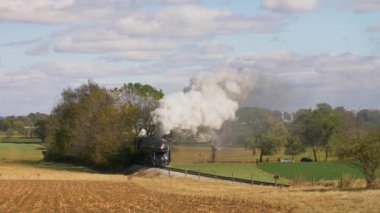 Antika buhar treni güneşli bir sonbahar gününde kırsal bölgelerde duman ve buhar üfleyerek seyahat ediyor.