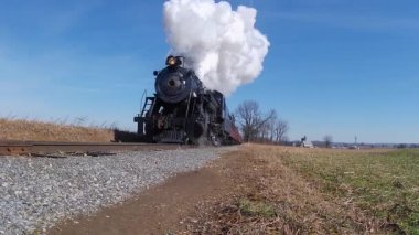Yenilenmiş bir buharlı lokomotifin düşük melek görüntüsü ve tren kırsal kesiminden takip ederek duman ve buhar üfleyen yolcu arabaları.