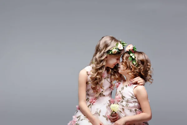 Two cute little girls - sisters in wreaths of fresh flowers on their heads sit and hug in a bright studio. Family and childhood friendship concept — Stock Photo, Image