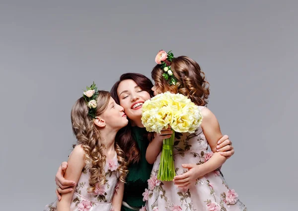 Beautiful happy family mom and two little cute girls in wreaths of fresh flowers and a large bouquet of tulips kiss, laugh and hug in the studio on a light background. — Stock Photo, Image