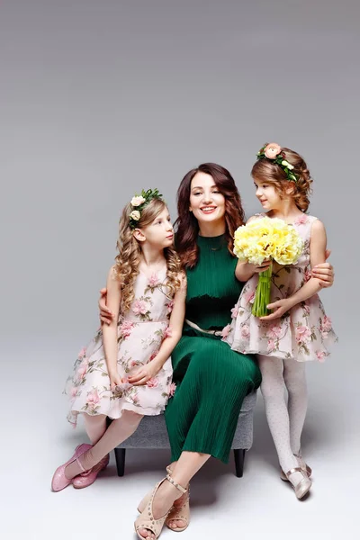 Beautiful happy family mom and two little cute girls in wreaths of fresh flowers and a large bouquet of tulips kiss, laugh and hug in the studio on a light background — Stock Photo, Image