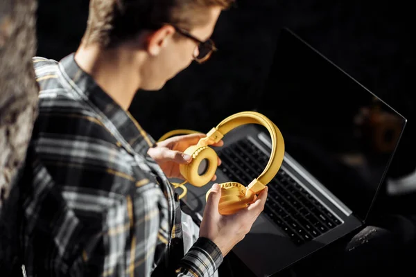 Foto vista posteriore di giovane studente di sesso maschile che tiene le cuffie gialle davanti al notebook o laptop mentre seduto sull'erba al parco . — Foto Stock