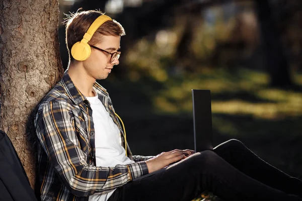 Knappe jonge mannelijke student in casual outfit met gele hoofdtelefoon met behulp van notebook of laptop terwijl zitten op het gras in het park. — Stockfoto