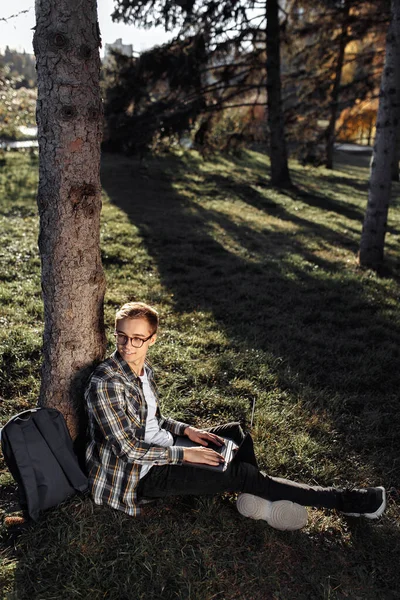 Jonge mannelijke student in casual outfit met behulp van notebook of laptop tijdens het zitten op het gras in het park. — Stockfoto