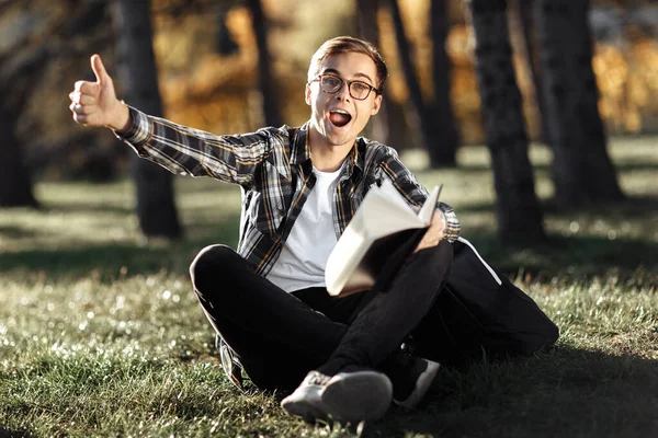 Excited young male student in casual outfit holding a book and show ok gesture while sitting on the grass at the park.