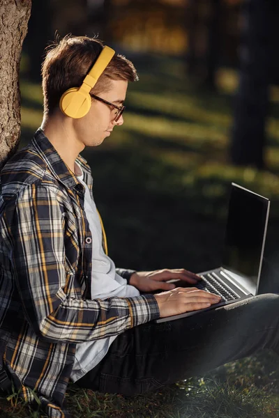 Mooie jonge mannelijke student in casual outfit met gele hoofdtelefoon met behulp van notebook of laptop terwijl zitten op het gras in het park. — Stockfoto