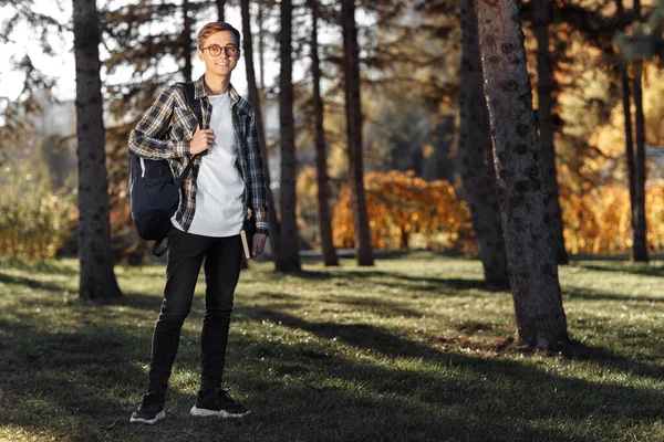 Full längd foto av glad ung manlig student med glasögon i casual outfit innehav ryggsäck och poserar i parken. — Stockfoto