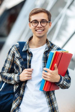 Neşeli genç bir erkek öğrenci, günlük kıyafetler içinde. Elinde parlak bir gülümsemeyle sokakta dikilirken klasörler tutuyor..