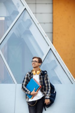 Sarı kulaklıklı, günlük kıyafetli parlak genç bir öğrenci. Elinde klasörlerle caddenin önünde dikiliyordu..