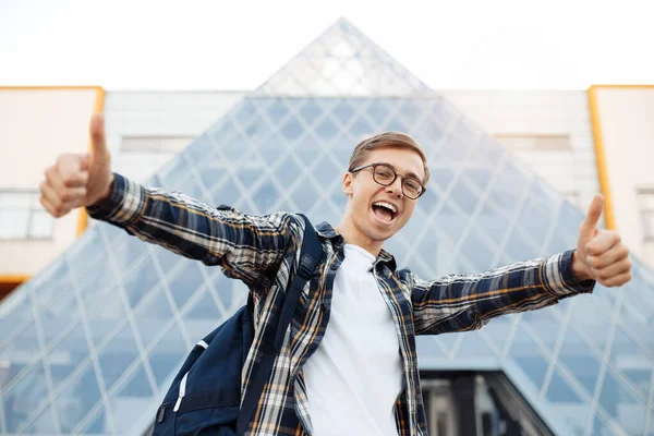 Vrolijke jonge mannelijke student in casual outfit met heldere glimlach holding show ok gebaar terwijl staan in de voorkant van driehoek gebouw op de straat. — Stockfoto