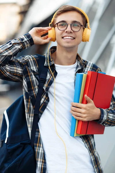 Close-up portret van jonge mannelijke student in casual outfit met gele hoofdtelefoon met mappen tijdens het luisteren naar muziek op straat. — Stockfoto
