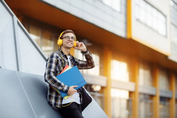 Knappe jonge mannelijke student in casual outfit met gele hoofdtelefoon met mappen tijdens het luisteren naar muziek op straat. — Stockfoto