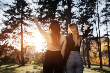 Happy two young twin sisters in casual outfit showing peace gesture at the park. Back view. Freedom Concept.