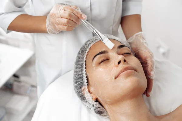 Kosmetolog stosujący krem na twarz kobiety pędzlem w salonie kosmetologii. — Zdjęcie stockowe