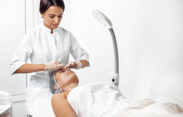 Cosmetoloog aanbrengen crème op vrouwelijk gezicht in cosmetologie salon. — Stockfoto