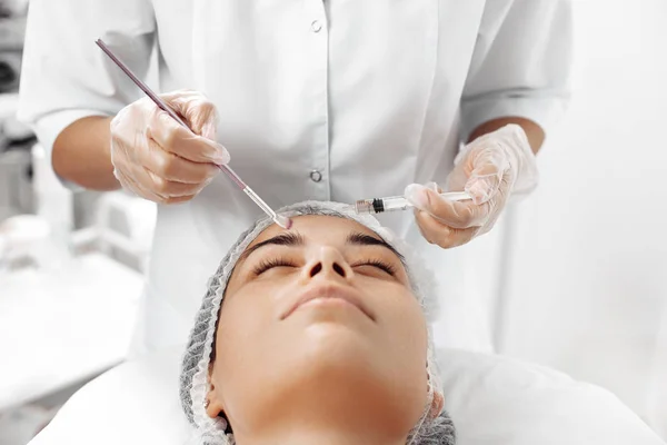 Kosmetolog stosujący krem na twarz kobiety pędzlem w salonie kosmetologii. — Zdjęcie stockowe