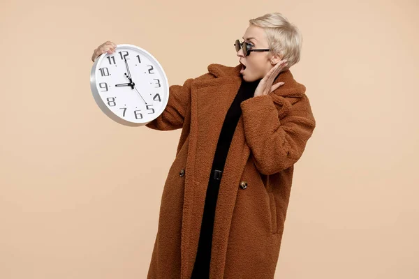 Schockierte junge Hipsterfrau mit blonden kurzen Haaren, Mantel und Sonnenbrille, die eine Uhr vor beigem Hintergrund hält. — Stockfoto