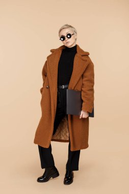 Sarı saçlı, ceketli, güneş gözlüklü, bej arka planda bilgisayarı olan son moda genç hippi kadın..