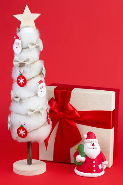 Gips bunte Weihnachtsmann auf der Geschenkbox und Weihnachtsbaum mit Dekorationen über rotem Hintergrund. — Stockfoto