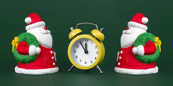 Zwei niedliche Gips bunte Weihnachtsmann und gelbe Uhr isoliert über grünem Hintergrund. Neujahrs- und Weihnachtskonzept. — Stockfoto