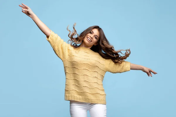 Happy young woman in yellow sweater dancing with hands up over blue background. — Stok fotoğraf