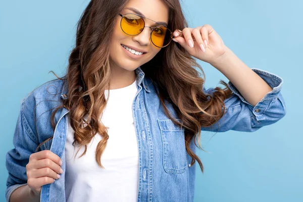 Boldog fiatal lány fényes mosollyal öltözött elegáns modern alkalmi ruhák és napfény pózol a kék háttér. — Stock Fotó