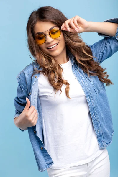 Boldog fiatal lány fényes mosollyal öltözött elegáns modern alkalmi ruhák és napfény pózol a kék háttér. — Stock Fotó