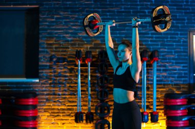 Formda genç bir kadın, spor salonunda halterle omuzlarına egzersiz yaptırıyor. Sağlık ve Spor kavramı.
