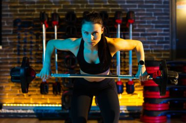 Formda genç bir kadın, spor salonunda halterle omuz ve sırt kaslarına egzersiz yaptırır. Sağlık ve Spor kavramı.