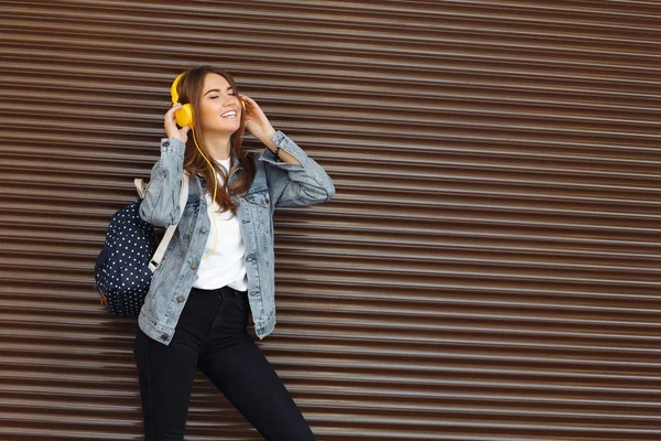 Friedlich lächelndes Mädchen mit braunen Haaren, das im Freien einen Rucksack und gelbe Kopfhörer trägt. junge Frau hört Musik. — Stockfoto