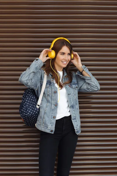 Hübsches lächelndes Mädchen mit braunen Haaren, das im Freien einen Rucksack und gelbe Kopfhörer trägt. junge Frau hört Musik. — Stockfoto