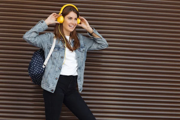 Glücklich lächelndes Mädchen mit braunen Haaren, das im Freien einen Rucksack und gelbe Kopfhörer trägt. junge Frau hört Musik. — Stockfoto