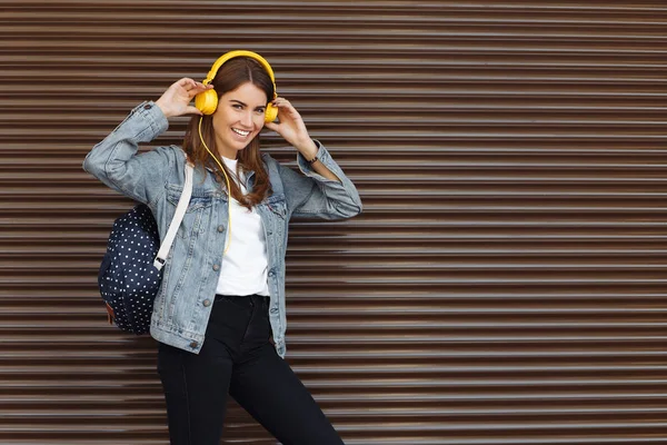 Trendy lächelndes Mädchen mit braunen Haaren trägt einen Rucksack und gelbe Kopfhörer im Freien. junge Frau hört Musik. — Stockfoto