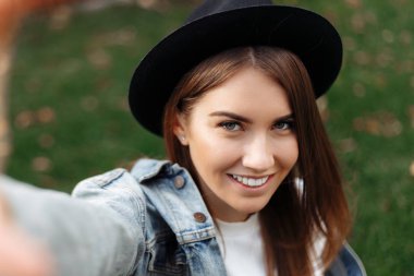 Kahverengi saçlı, şapkalı, sırt çantalı modern hippi kız parkta akıllı telefonlu selfie çekerken....