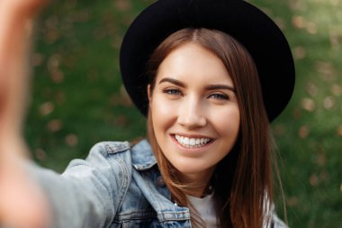 Kahverengi saçlı, şapkalı ve sırt çantalı neşeli hippi kız parkta akıllı telefonlu selfie çekerken....